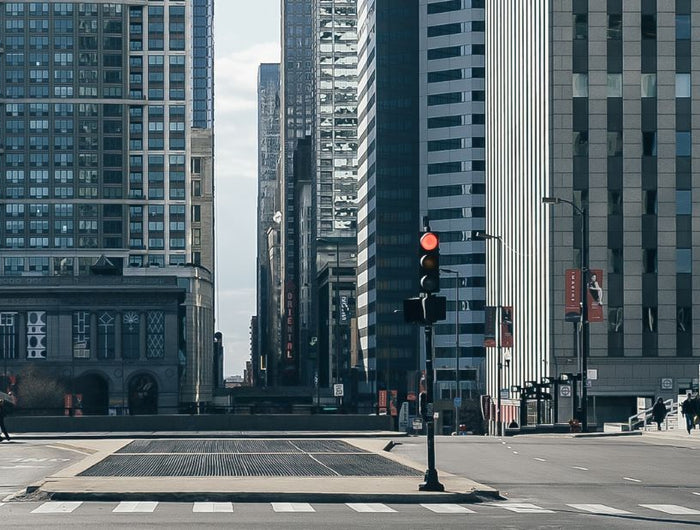 Breite Stadtstraße, flankiert von hohen Gebäuden, meist aus Glas und Beton. Nur wenige Fußgänger überqueren die Straße in der Nähe eines glasüberdachten Transiteingangs. Ampeln und Fußgängerüberwege sind sichtbar. Die Atmosphäre erscheint ruhig, mit sanftem Tageslicht und wenig Verkehr.